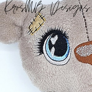 Könnte beinhalten: Nahaufnahme eines Plüschtierkopfes mit gestickten Details. Das Spielzeug hat eine hellgraue, flauschige Textur, ein großes Auge mit blauer und schwarzer Stickerei und einen kleinen, goldfarbenen Aufnäher, der an der Seite angenäht ist. Der Text "KrosMB Designs" ist kursiv geschrieben.