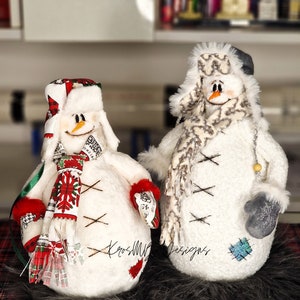 May include: Two white snowman figurines with red and white scarves and hats. The snowman on the left has a red and white snowflake pattern on its scarf. The snowman on the right has a gray and white scarf. Both snowmen have black eyes and a carrot nose. The snowmen are sitting on a black and red plaid surface.
