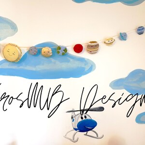 May include: A white wall with a blue and white painted cloud design. A string of felt planets and a rocket ship hangs from the clouds. The text "KrosMUB Designs" is written in black cursive on the wall. A blue helicopter is painted on the wall below the text.