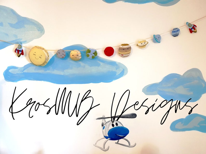 May include: A white wall with a blue and white painted cloud design. A string of felt planets and a rocket ship hangs from the clouds. The text "KrosMUB Designs" is written in black cursive on the wall. A blue helicopter is painted on the wall below the text.