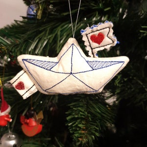 May include: White fabric ornament shaped like a paper boat with blue stitching and a red heart on a white fabric flag attached to the top.