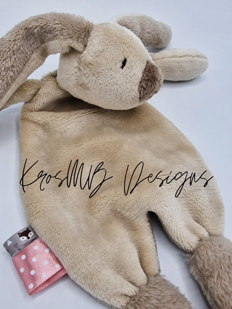 May include: A tan plush bunny lovey with a brown nose and a pink and white polka dot ribbon attached. The lovey has a small fox design on the ribbon. The text "KrossMB Designs" is visible on the lovey.