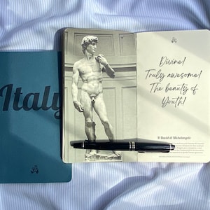 Può includere: Un quaderno blu con la parola "Italia" in lettere bianche. Il quaderno è aperto su una pagina con un'immagine in bianco e nero della statua di David di Michelangelo. La pagina contiene anche un testo scritto a mano che dice "Divino! Davvero fantastico! La bellezza della giovinezza!" e "Il David di Michelangelo".