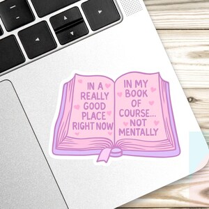 May include: A pink sticker in the shape of an open book with the text "IN A REALLY GOOD PLACE RIGHT NOW" on the left page and "IN MY BOOK OF COURSE... NOT MENTALLY" on the right. The sticker is on a laptop.