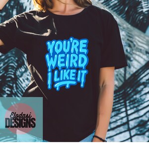 Peut inclure: T-shirt noir avec le texte "YOU'RE WEIRD I LIKE IT" en lettres bleues à bulles. Le t-shirt présente également le logo "Elodasi DESIGNS" dans un encadré turquoise. Le t-shirt est de style col rond.