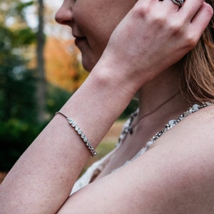 Peut inclure: Une femme portant une robe blanche avec un bracelet argenté et une bague argentée avec un gros diamant. Elle se touche les cheveux avec sa main droite.