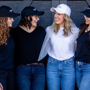 以下が含まれることがあります： 4人の友達が写真にポーズをとっています。3人は「Squad」と刺繍された黒い野球帽をかぶり、1人は「Bride」と刺繍された白い野球帽をかぶっています。花嫁は白いTシャツと青いジーンズを着用し、頭にベールをかぶっています。