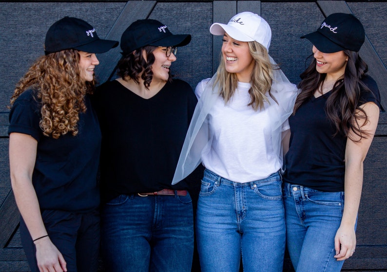 以下が含まれることがあります： 4人の友達が写真にポーズをとっています。3人は「Squad」と刺繍された黒い野球帽をかぶり、1人は「Bride」と刺繍された白い野球帽をかぶっています。花嫁は白いTシャツと青いジーンズを着用し、頭にベールをかぶっています。