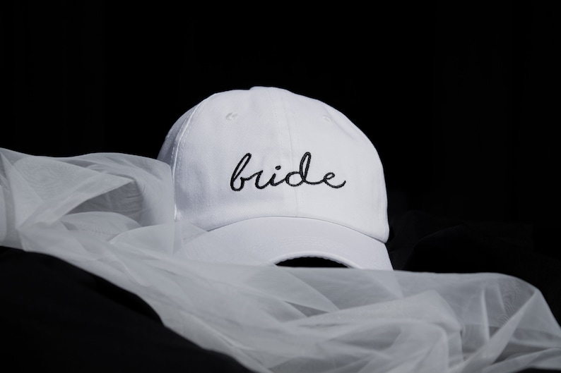 以下が含まれることがあります： 白い野球帽に、黒い筆記体で「bride」と刺繍されています。帽子は白いチュール生地の上に置かれています。