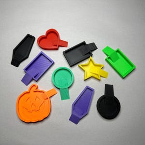 May include: A set of ten 3D printed shapes in various colors, including a heart, a star, a pumpkin, a circle, and a rectangle. The shapes are all small and have a flat back with a small tab for attaching them to a surface.