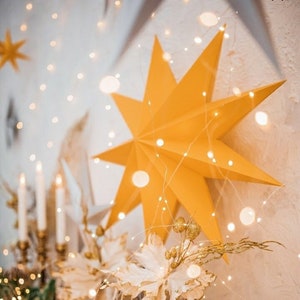 Puede incluir: Una estrella de papel amarilla con una cadena de luces blancas colgando de ella. La estrella está colgada en una pared blanca con una guirnalda verde y una jaula de pájaros blanca en primer plano.
