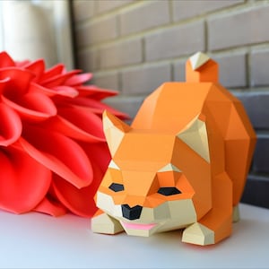 Puede incluir: Un modelo de papel de un perro Shiba Inu naranja y tostado. El perro está sentado sobre una superficie blanca con una flor roja en el fondo.