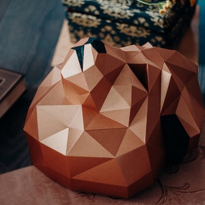 Puede incluir: Una escultura de cabeza de oso geométrica de papel marrón con detalles negros. La escultura está hecha de papel doblado y tiene un diseño tridimensional.