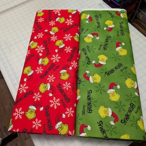 May include: Two fabric bolts, one red and one green, featuring a repeating pattern of the Grinch character, snowflakes, and the text "Merry Grinchmas." The red fabric has a white snowflake design.