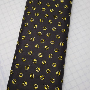 Puede incluir: Tela negra con un patr&oacute;n repetido de logotipos de Batman amarillos. La tela tiene el texto "Camelot Fabrics. camelotfabrics.com Not intended for children's sleepwear."