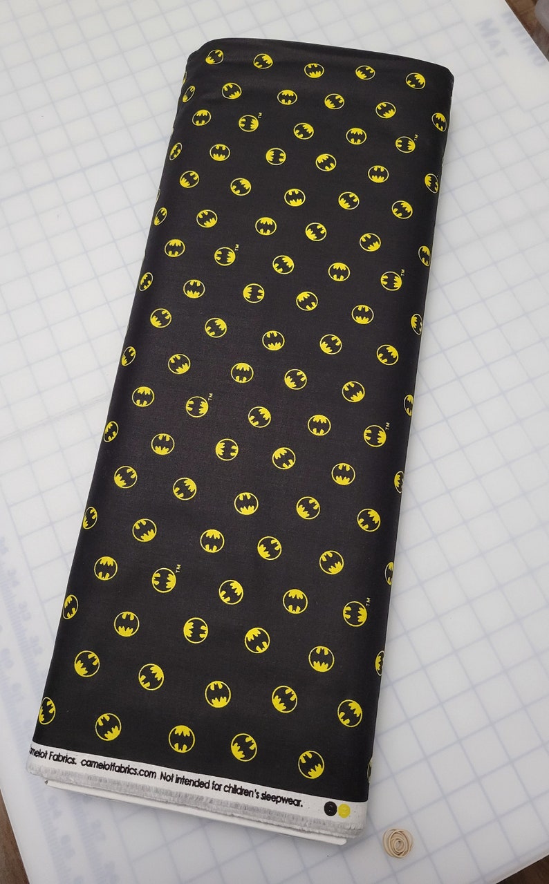 Puede incluir: Tela negra con un patr&oacute;n repetido de logotipos de Batman amarillos. La tela tiene el texto "Camelot Fabrics. camelotfabrics.com Not intended for children's sleepwear."