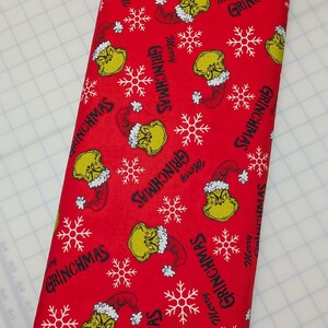 May include: A roll of red fabric featuring a repeating pattern of the Grinch's face, snowflakes, and the text "Merry Grinchmas" in white. The fabric is suitable for holiday-themed crafts and sewing projects.