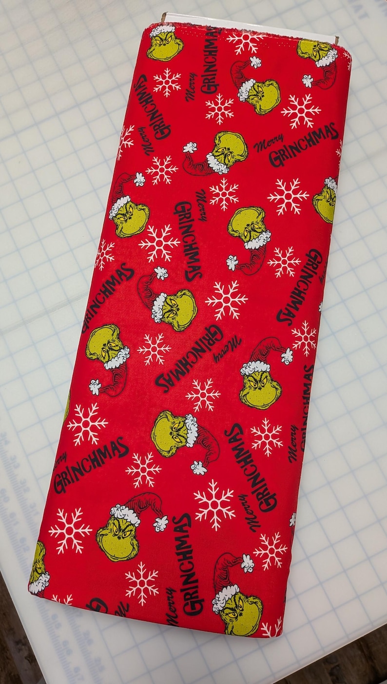 May include: A roll of red fabric featuring a repeating pattern of the Grinch's face, snowflakes, and the text "Merry Grinchmas" in white. The fabric is suitable for holiday-themed crafts and sewing projects.