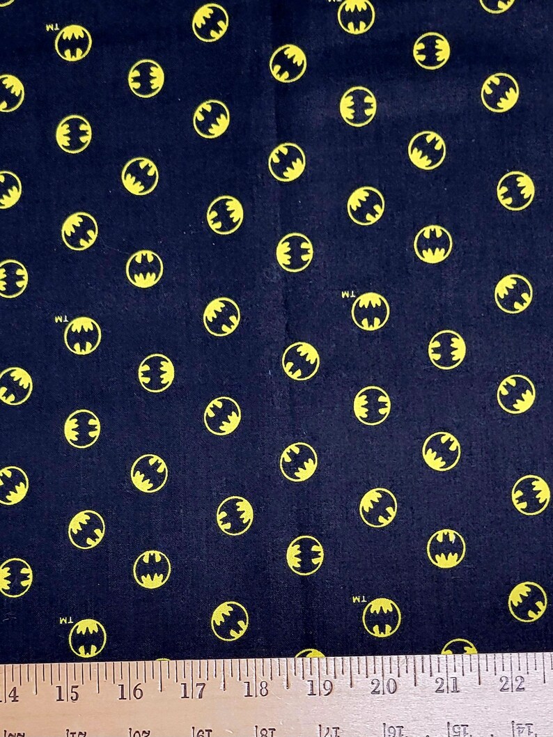 Puede incluir: Tela negra con un patr&oacute;n repetido de logotipos de Batman amarillos. La tela se mide con una regla en pulgadas.