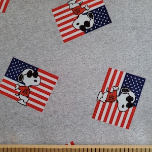 May include: Gray fabric with a repeating pattern of a cartoon dog wearing sunglasses and a red shirt standing on an American flag. The dog is wearing a red shirt with the text "USA" on it. The fabric is measured with a wooden ruler.