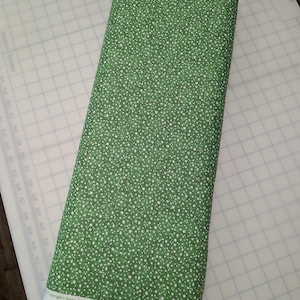 May include: A tightly rolled bolt of green fabric with a white polka dot pattern. The fabric is on a white cutting mat. The text "Copyright Susan Winger Licensed to Wilmington Prints. All Rights Reserved" is visible.