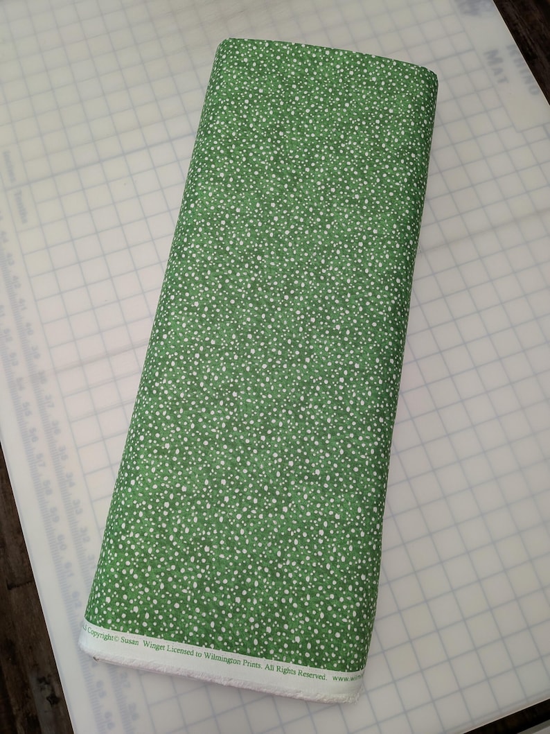 May include: A tightly rolled bolt of green fabric with a white polka dot pattern. The fabric is on a white cutting mat. The text "Copyright Susan Winger Licensed to Wilmington Prints. All Rights Reserved" is visible.