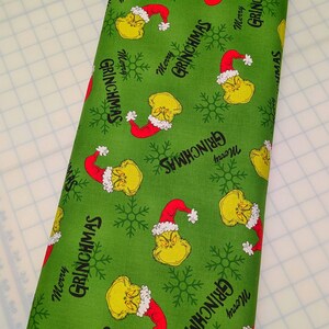 May include: Green fabric roll with a repeating pattern of a yellow character wearing a red Santa hat, snowflakes, and the text "Merry Grinchmas". The fabric is suitable for holiday-themed crafts and sewing projects.