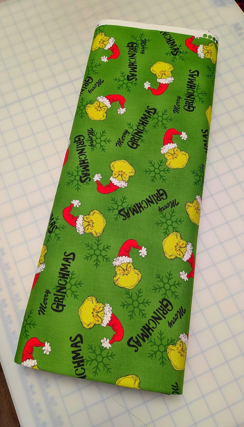 May include: Green fabric roll with a repeating pattern of a yellow character wearing a red Santa hat, snowflakes, and the text "Merry Grinchmas". The fabric is suitable for holiday-themed crafts and sewing projects.