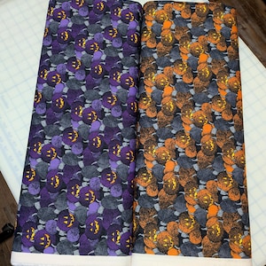 May include: Two folded bolts of fabric featuring a repeating pattern of jack-o'-lanterns and textured gray circles. One bolt displays purple pumpkins with yellow faces, while the other features orange pumpkins with yellow faces on a dark background.