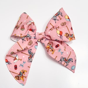 Puede incluir: Un lazo rosa con un estampado de carrusel con cebras, leones y otros animales. El lazo está hecho de tela y tiene un respaldo blanco.