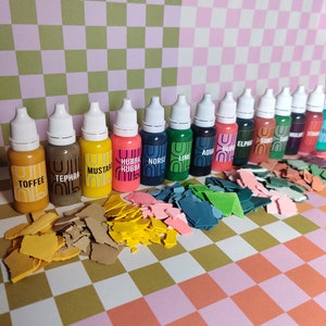 May include: A set of 14 small bottles of paint with white lids. The bottles are arranged in a row on a checkered surface. Each bottle has a different color of paint and a label with a name. The colors include: Toffee, Tephra, Mustard, Hubba Hubba, Norse, Lime, Aqua, Bliss, Elphine, Suns, Sails, Mullie, Straw, and Sea.