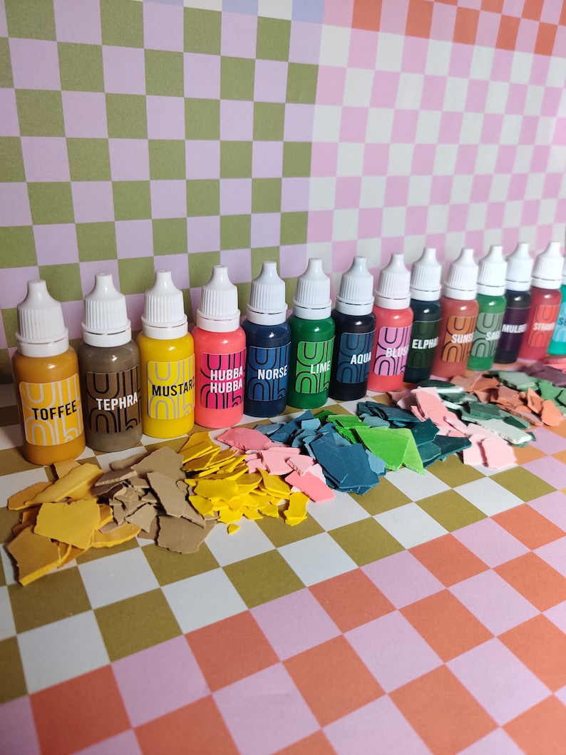 May include: A set of 14 small bottles of paint with white lids. The bottles are arranged in a row on a checkered surface. Each bottle has a different color of paint and a label with a name. The colors include: Toffee, Tephra, Mustard, Hubba Hubba, Norse, Lime, Aqua, Bliss, Elphine, Suns, Sails, Mullie, Straw, and Sea.