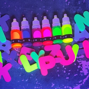 May include: Six small bottles of neon colored paint, with labels that say "ORAN", "BLON", "PON", "PION", "GION", and "Y". The bottles are arranged in a row on a dark surface, surrounded by neon colored letters.