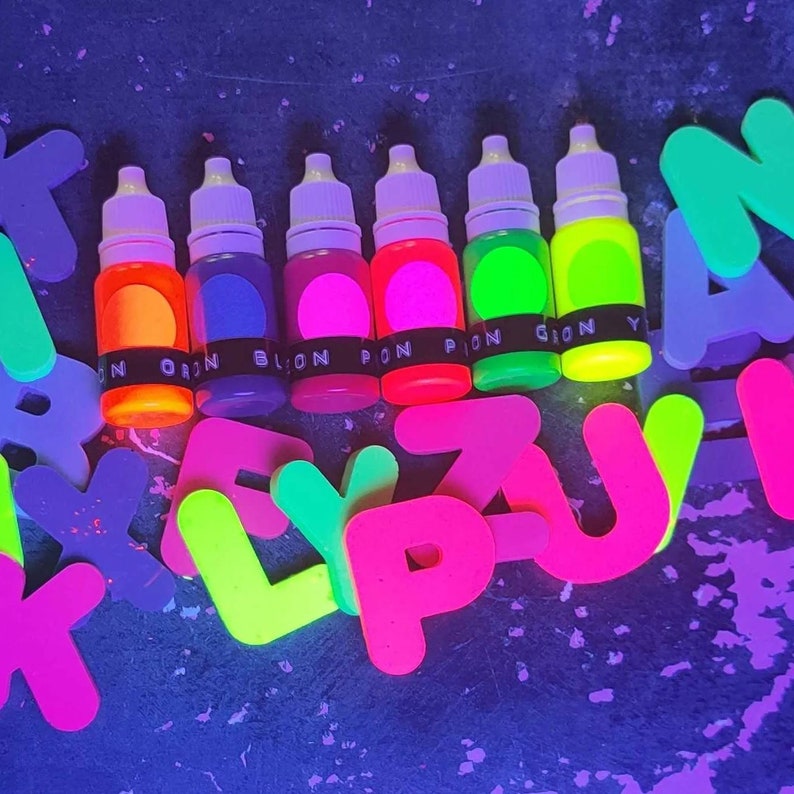 May include: Six small bottles of neon colored paint, with labels that say "ORAN", "BLON", "PON", "PION", "GION", and "Y". The bottles are arranged in a row on a dark surface, surrounded by neon colored letters.