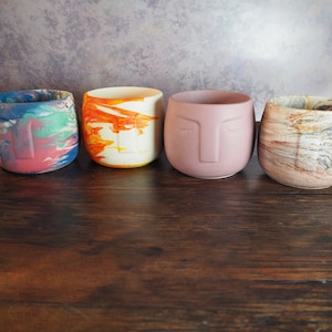 May include: Four small, round, concrete planters with different marbled patterns. The planters are in a row on a wooden surface. The planters are all different colors, including blue, orange, pink, and brown.