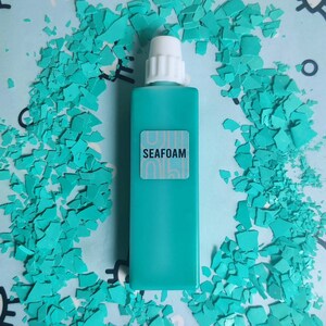 May include: A turquoise blue bottle with a white cap and a white label that says "SEAFOAM". The bottle is sitting on a surface covered in turquoise blue confetti.