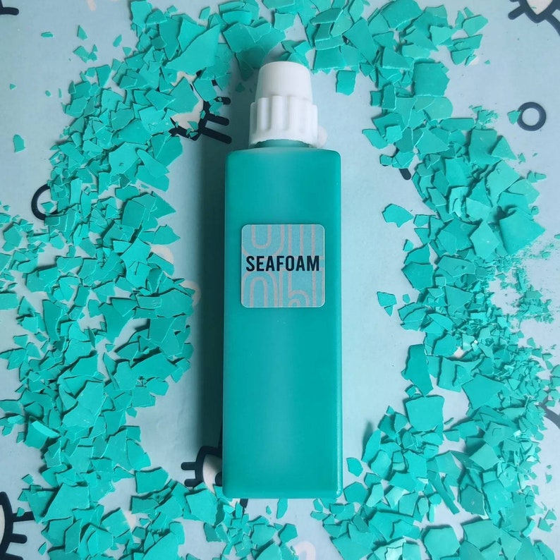May include: A turquoise blue bottle with a white cap and a white label that says "SEAFOAM". The bottle is sitting on a surface covered in turquoise blue confetti.
