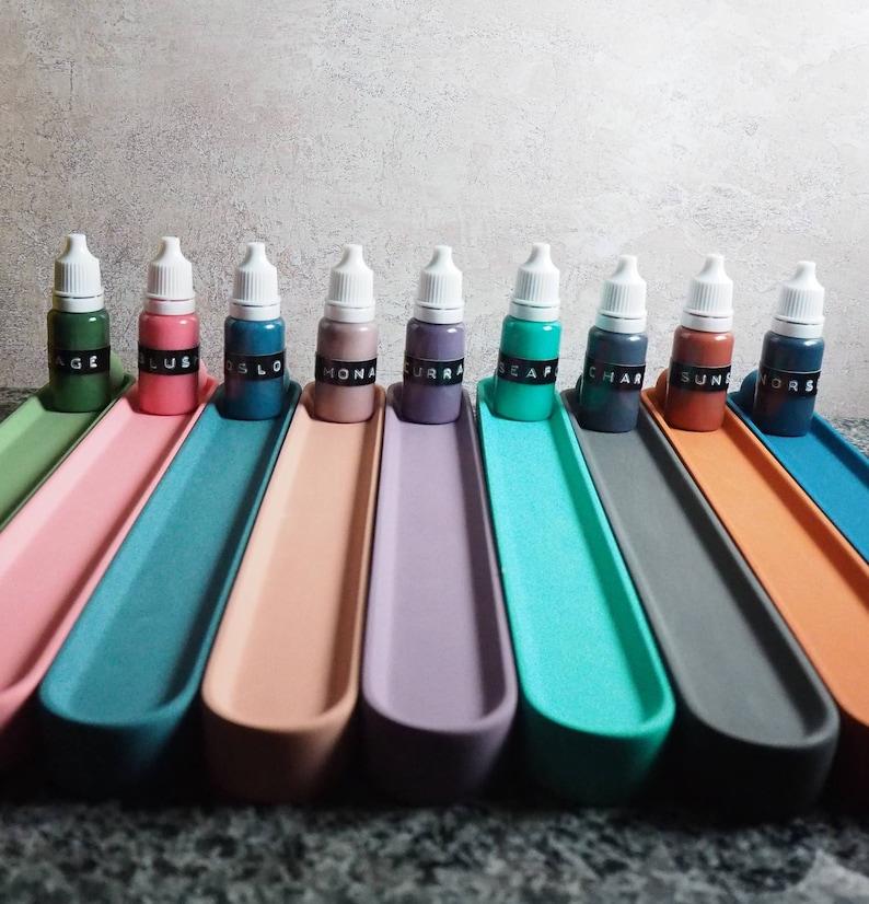 May include: Ten small bottles of liquid paint, each with a different color, are lined up on a shelf. The bottles are labeled with names like "Sage", "Oslo", "Mocha", "Curra", "Seafoam", "Char", "Sun", and "Nors".
