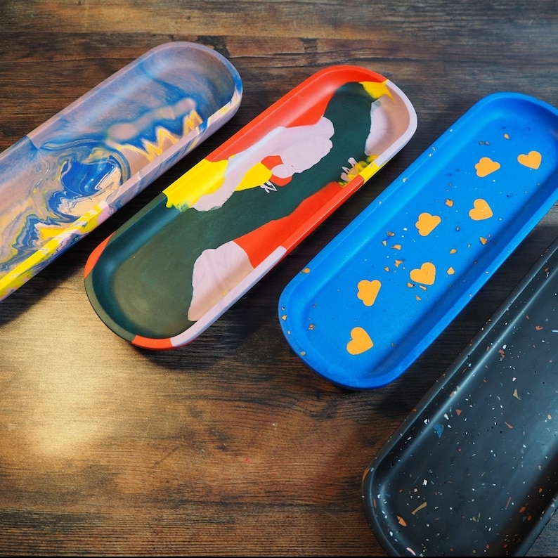 May include: Four colorful ceramic trays with different patterns. The trays are oval shaped and have a smooth, glossy finish. The patterns include a blue and pink swirl, a red, green, yellow, and pink abstract design, a blue tray with gold flecks and yellow hearts, and a black tray with white flecks.