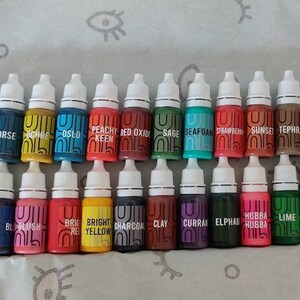 May include: A collection of 30 small bottles of paint, arranged in two rows. The bottles are labeled with names like "Aqua", "Black", "Blush", "Bright Yellow", "Charcoal", "Clay", "Currant", "Elphas", "Hubba Hubba", "Lime", "Lola", "Midnight", "Mona", "Oslo", "Peachy Keen", "Red Oxide", "Sage", "Seafoam", "Strawberry", "Sunset", "Tephra", "Toffee", "Truffle", "White", "Beret", "Mustard", "Norse", "Ochre".