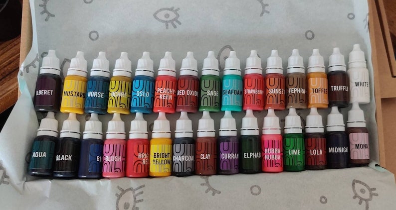 May include: A collection of 30 small bottles of paint, arranged in two rows. The bottles are labeled with names like "Aqua", "Black", "Blush", "Bright Yellow", "Charcoal", "Clay", "Currant", "Elphas", "Hubba Hubba", "Lime", "Lola", "Midnight", "Mona", "Oslo", "Peachy Keen", "Red Oxide", "Sage", "Seafoam", "Strawberry", "Sunset", "Tephra", "Toffee", "Truffle", "White", "Beret", "Mustard", "Norse", "Ochre".