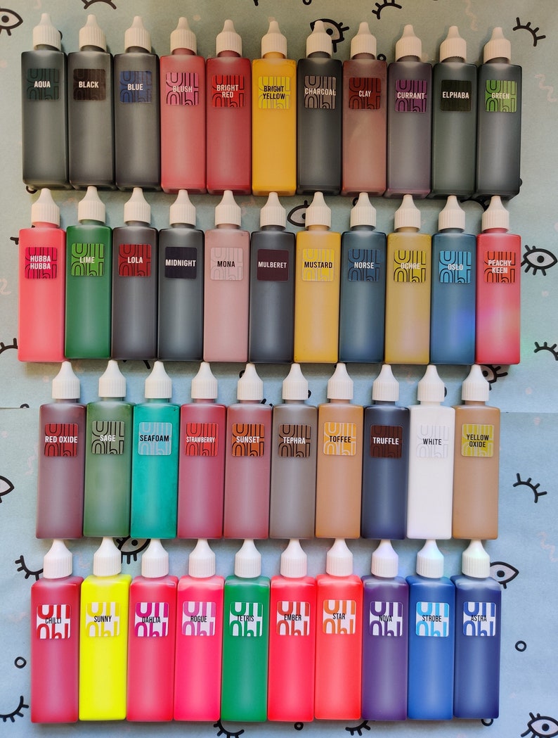 May include: A collection of 40 small glass bottles with white caps, each filled with a different colored liquid. The bottles are arranged in four rows of ten, and each bottle has a label with a name printed on it. The names include colors like Aqua, Black, Blue, Blush, Bright Red, Bright Yellow, Charcoal, Clay, Currant, Elphaba, Green, Hubba Hubba, Lime, Lola, Midnight, Mona, Mulberet, Mustard, Norse, Peachy Red, Red Oxide, Sage, Seafoam, Strawberry, Sunset, Tephra, Toffee, Truffle, White, Yellow Oxide, Chili, Sunny, Rogue, Tetris, Ember, Star, Nova, Strobe, and Astra.