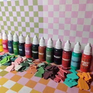 May include: A set of 12 small bottles of paint with dropper tops. The bottles are arranged in a row on a checkered surface. The paint colors are:  ard ba, bba orse, ume, qua, kush, phaba, sunset, sage, ulbere, strawberry, seafoam, peachy keen.