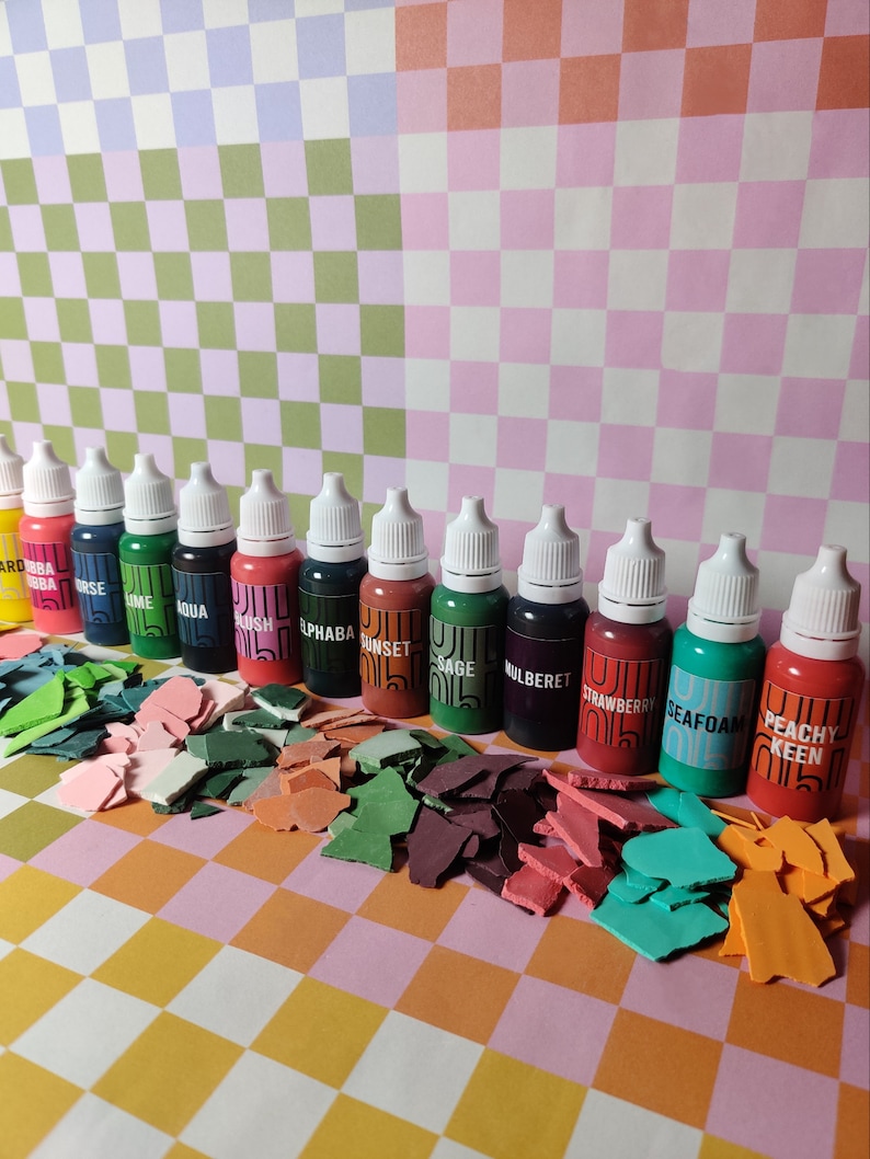 May include: A set of 12 small bottles of paint with dropper tops. The bottles are arranged in a row on a checkered surface. The paint colors are:  ard ba, bba orse, ume, qua, kush, phaba, sunset, sage, ulbere, strawberry, seafoam, peachy keen.