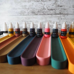 May include: A set of 14 small bottles of paint, each with a white cap and a black label with a color name. The bottles are arranged in a row on a set of colorful, curved trays.