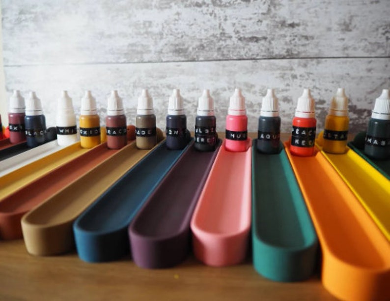 May include: A set of 14 small bottles of paint, each with a white cap and a black label with a color name. The bottles are arranged in a row on a set of colorful, curved trays.