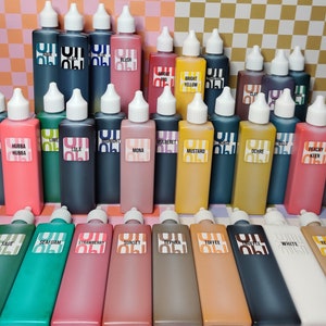 May include: A collection of 24 small glass bottles with colorful liquid inside. The bottles are arranged in four rows of six bottles each. The bottles are labeled with different names, such as "Sage", "Seafoam", "Strawberry", "Sunset", "Tephera", "Toffee", "White", and "Yellow Oxide".