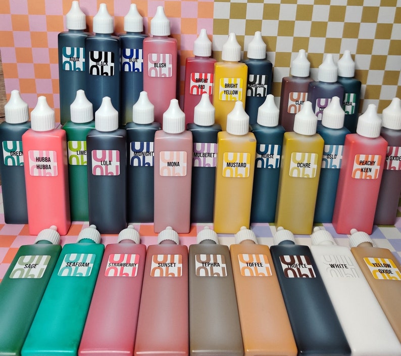 May include: A collection of 24 small glass bottles with colorful liquid inside. The bottles are arranged in four rows of six bottles each. The bottles are labeled with different names, such as "Sage", "Seafoam", "Strawberry", "Sunset", "Tephera", "Toffee", "White", and "Yellow Oxide".