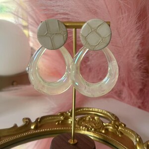 May include: A pair of iridescent acrylic hoop earrings with a light gray, textured top. The earrings are displayed on a gold-colored stand with a wooden base. The background features pink feathers and a gold-framed mirror.