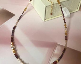 Peut inclure: Un collier de perles avec un pendentif doré. Le collier présente de petites perles rouge foncé, jaunes et transparentes. Le pendentif doré a une surface texturée. Le collier est présenté sur une surface rose à côté d'une boîte à bijoux blanche.